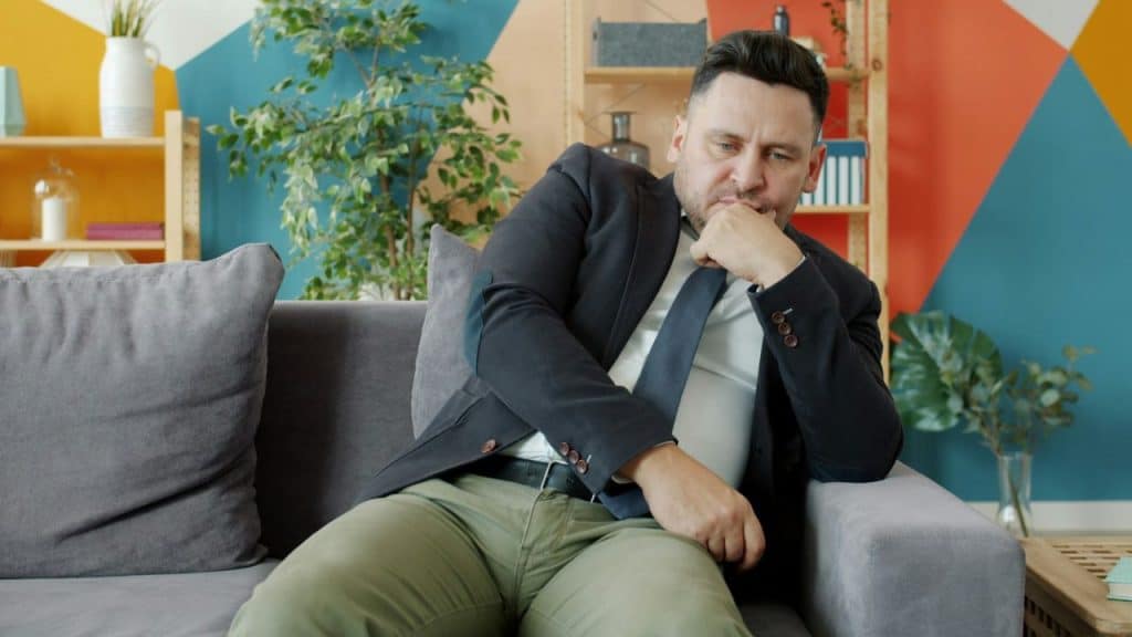 A man in a suit sits on a gray couch with his hand on his chin.