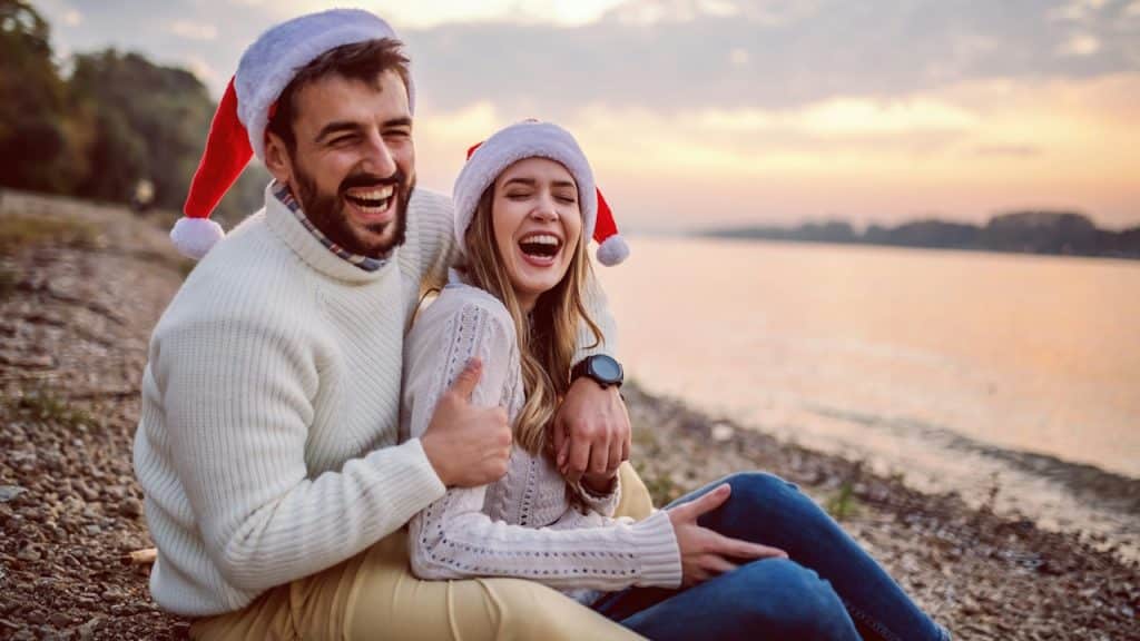 A couple with Christmas caps are laughing hard while sitting on a beach.