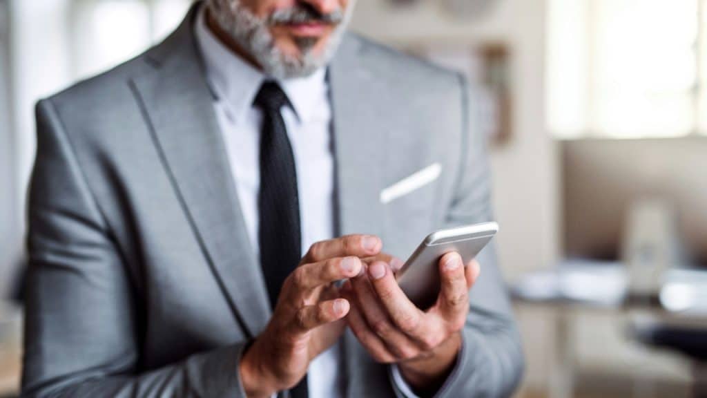 A man in a grey suit and tie holds a silver smartphone with both hands.