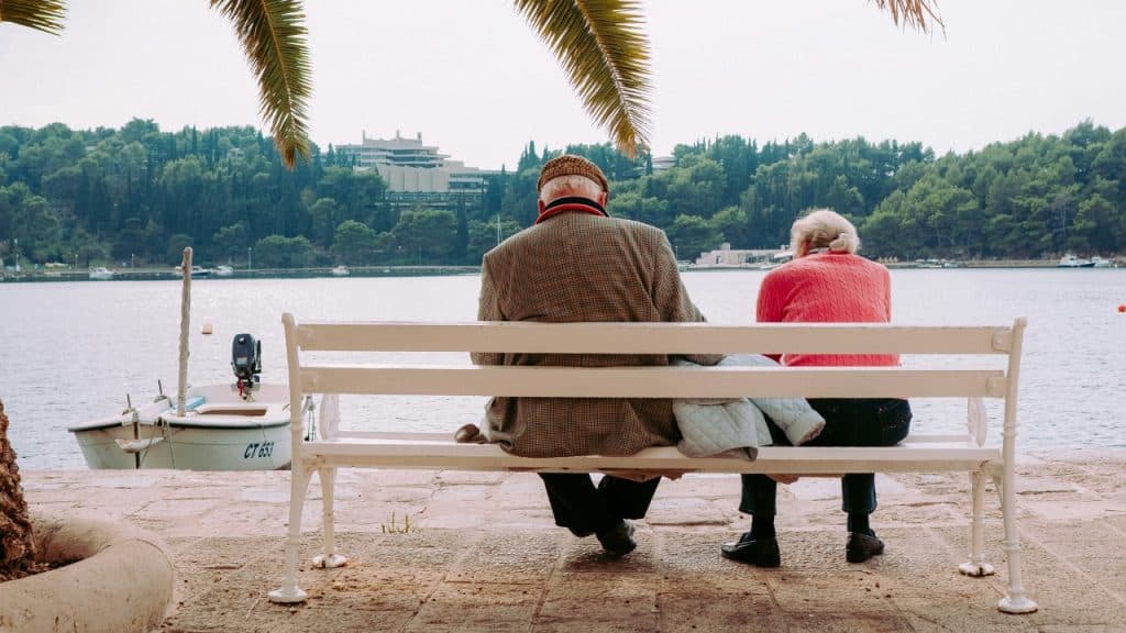 Two people sit on a white bench facing a body of water with a boat.