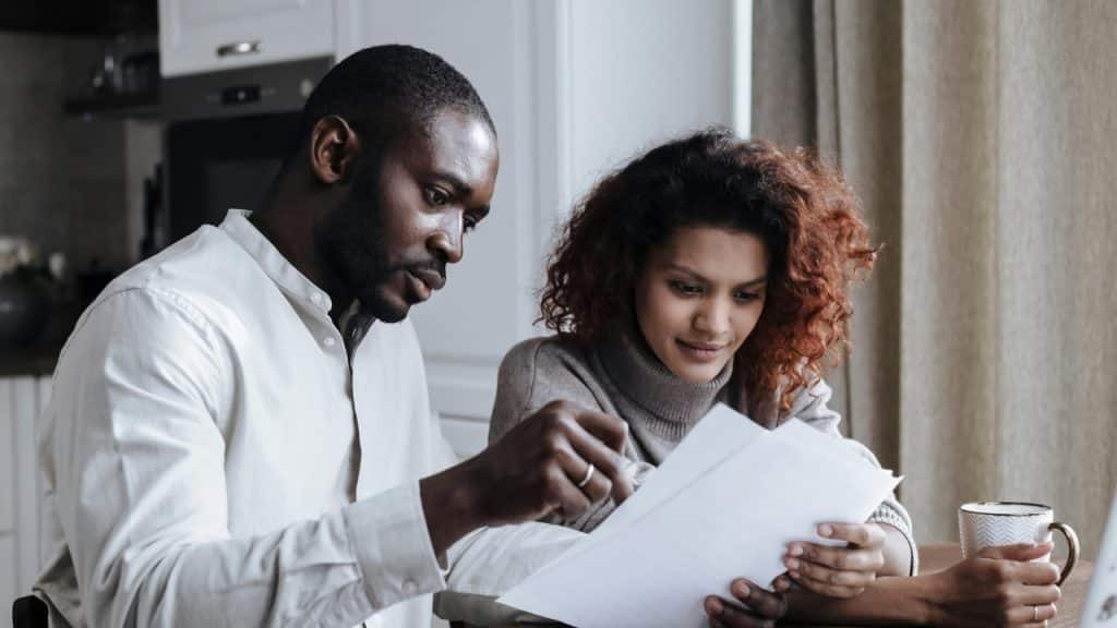 A man and woman sit at a wooden table together, looking intently at white papers.