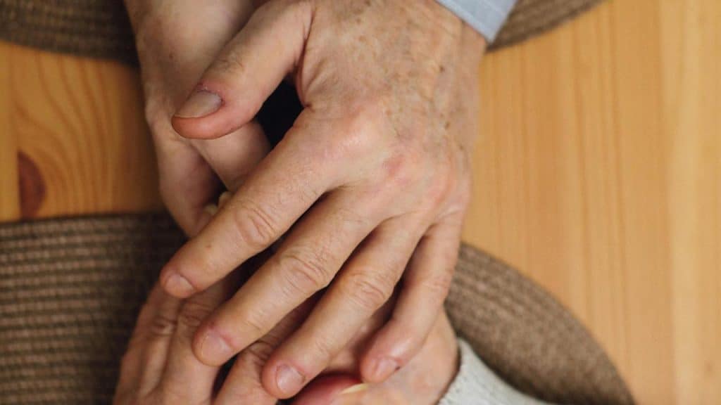 A pair of elderly hands gently holding each other on a wooden table.
