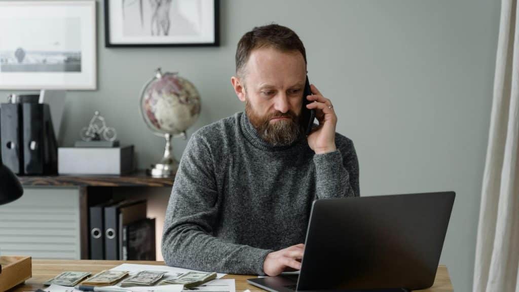 A man talking on the phone while working on a laptop with cash on the desk.