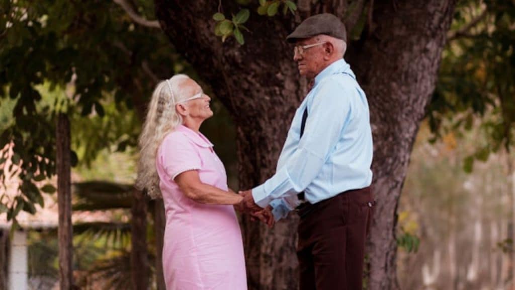 An older man and woman hold hands outdoors while standing under a large tree.