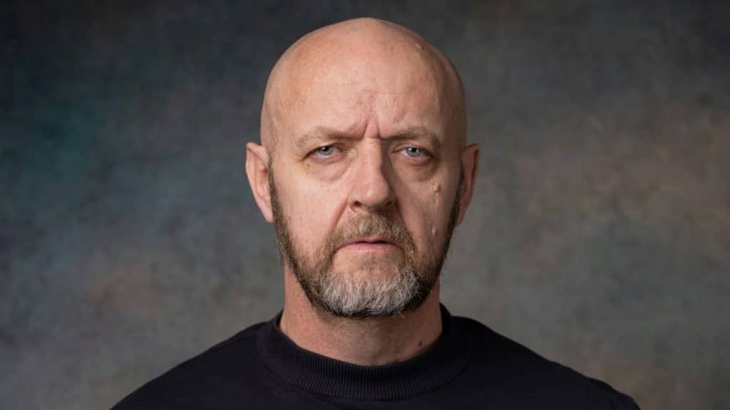 Close-up portrait of a bald man with a graying beard against a textured gray background.