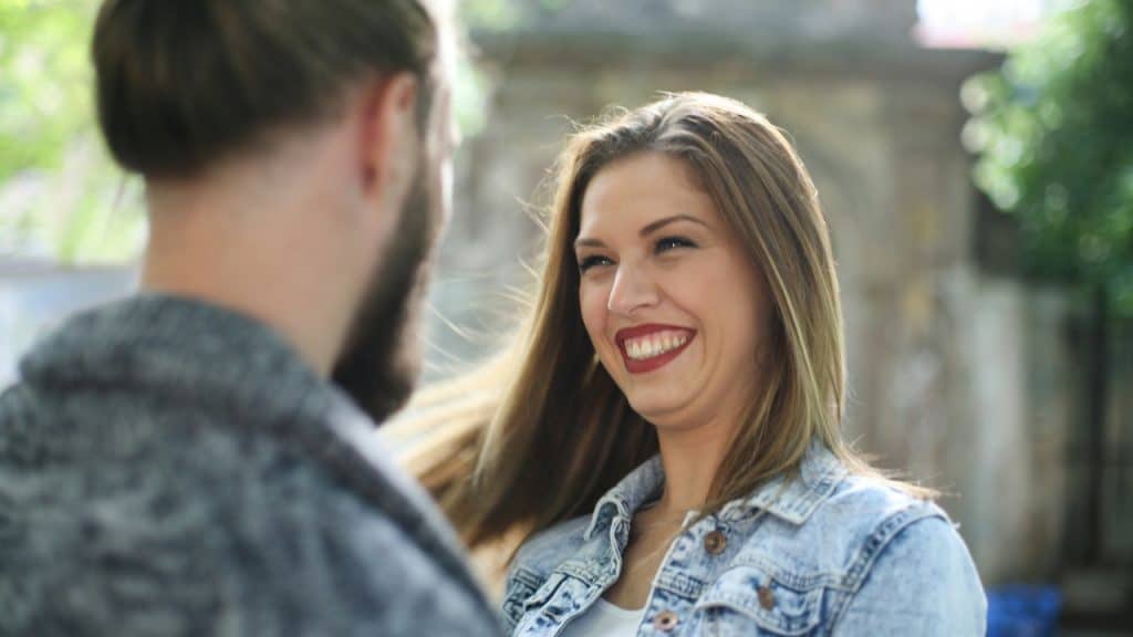 A woman in a denim jacket laughs while looking at a man with long hair.