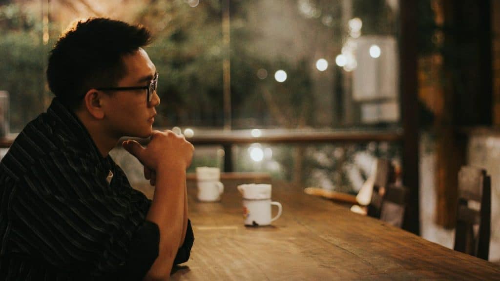 A man wearing glasses sits alone at a long wooden table with his hand on chin.