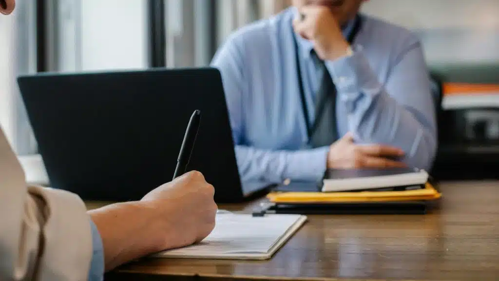 A person's hand writes on a document with a blurred person sitting in the background.