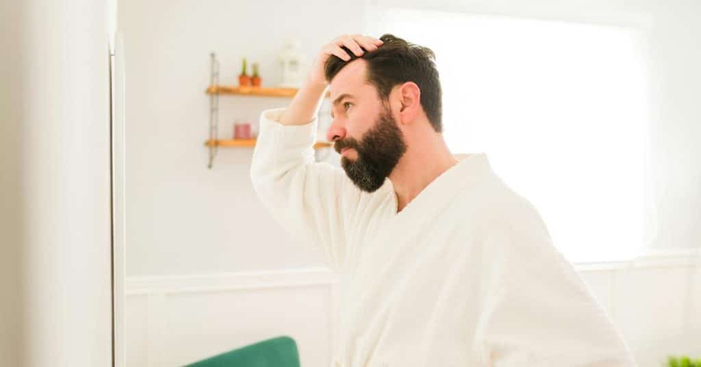 A bearded man in a robe touching his hair while looking in a mirror.