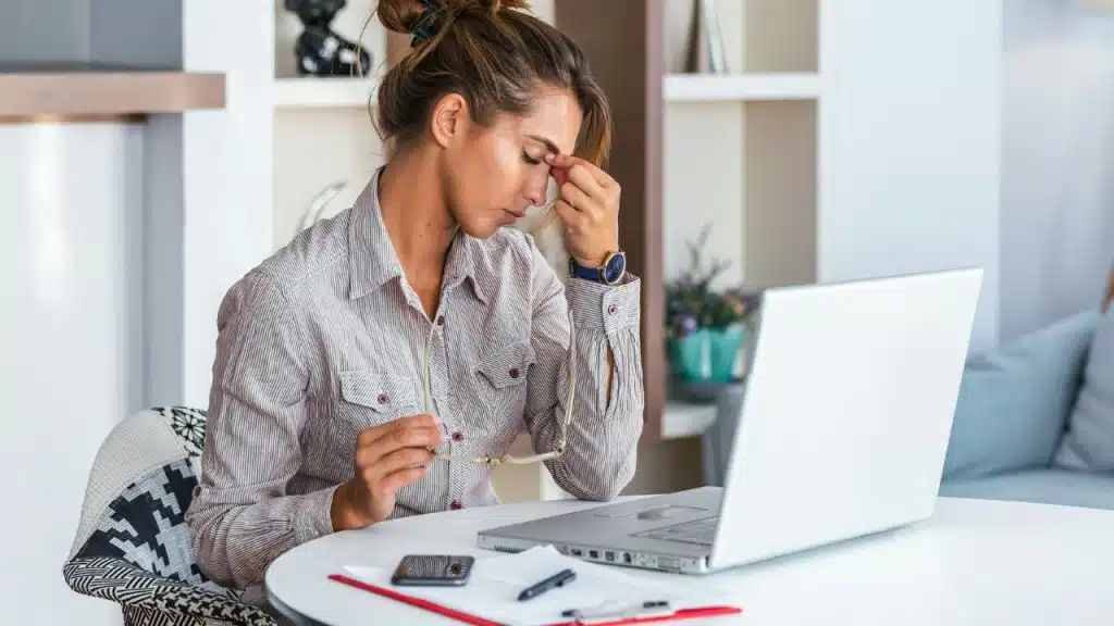 A woman sits at a table with a laptop, holding her glasses and pinching her nose.