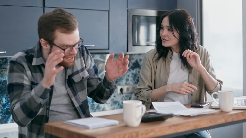 A man in glasses gestures toward papers while a woman watches him from the side.