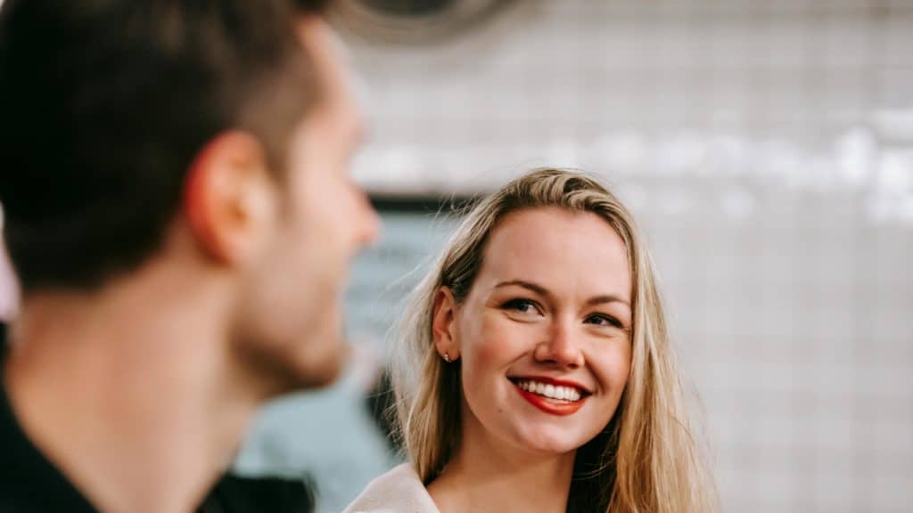 A smiling woman with red lipstick looks toward a man whose face is partially blurred.