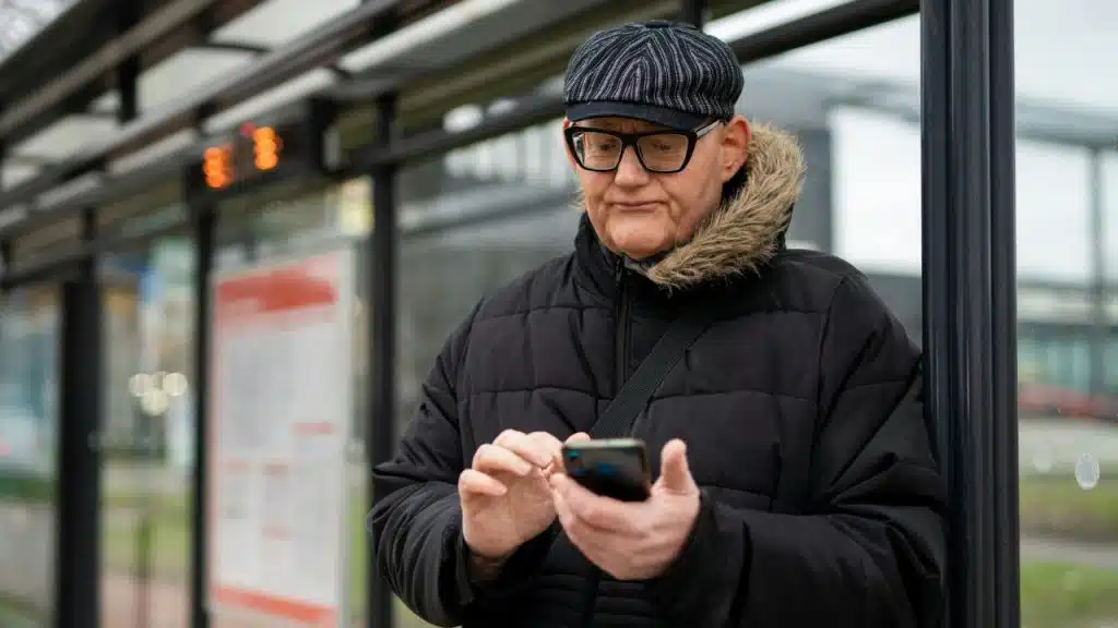 A man wearing a flat cap and glasses uses a smartphone while leaning against a post.