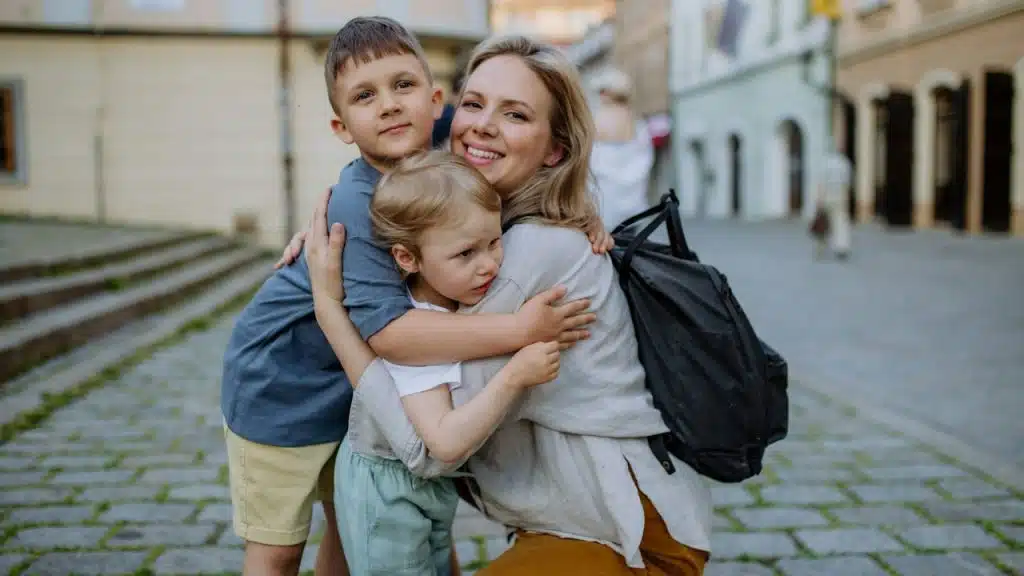 A young mother and little children smiling confidently and hugging each other while standing at a street.