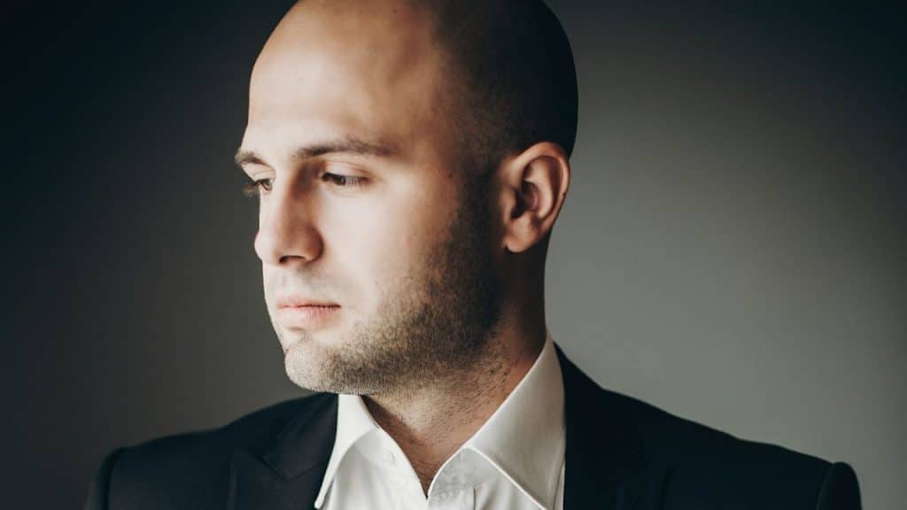 A pensive, bald man in a black suit and white shirt looks down and away from the camera.