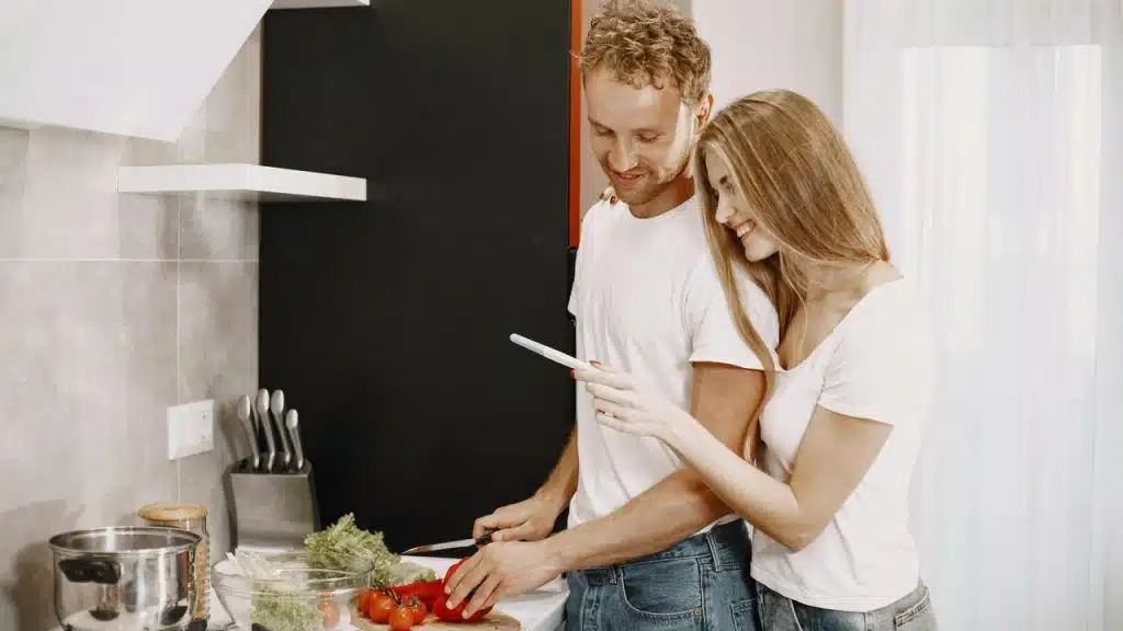 A couple cooking together in a kitchen while looking at a phone.