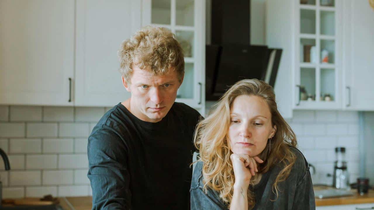 A serious couple standing together in a kitchen, looking worried.