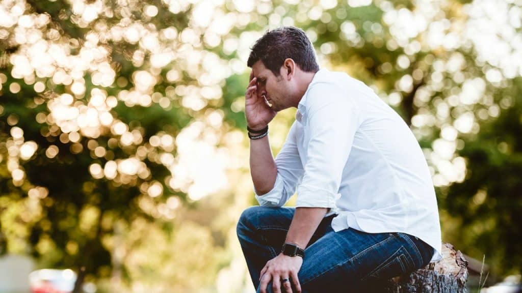 A man in a white shirt sits outside on a stump with his hand on his head.