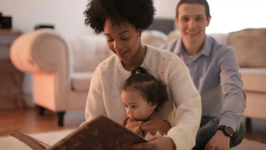 A woman reads a book to a child while a man smiles in the background.