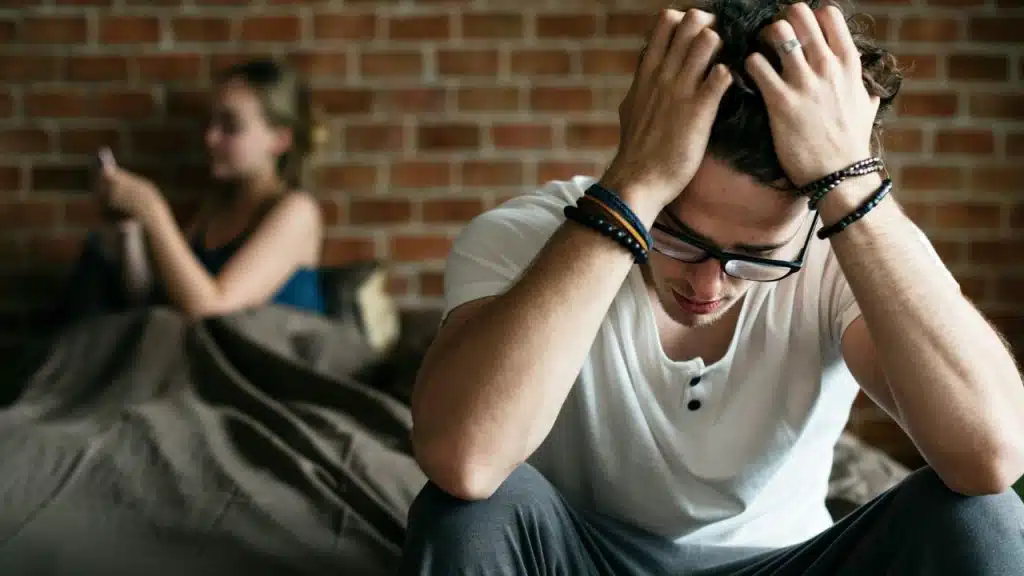 A man holds his head in his hands while a woman uses a phone behind.