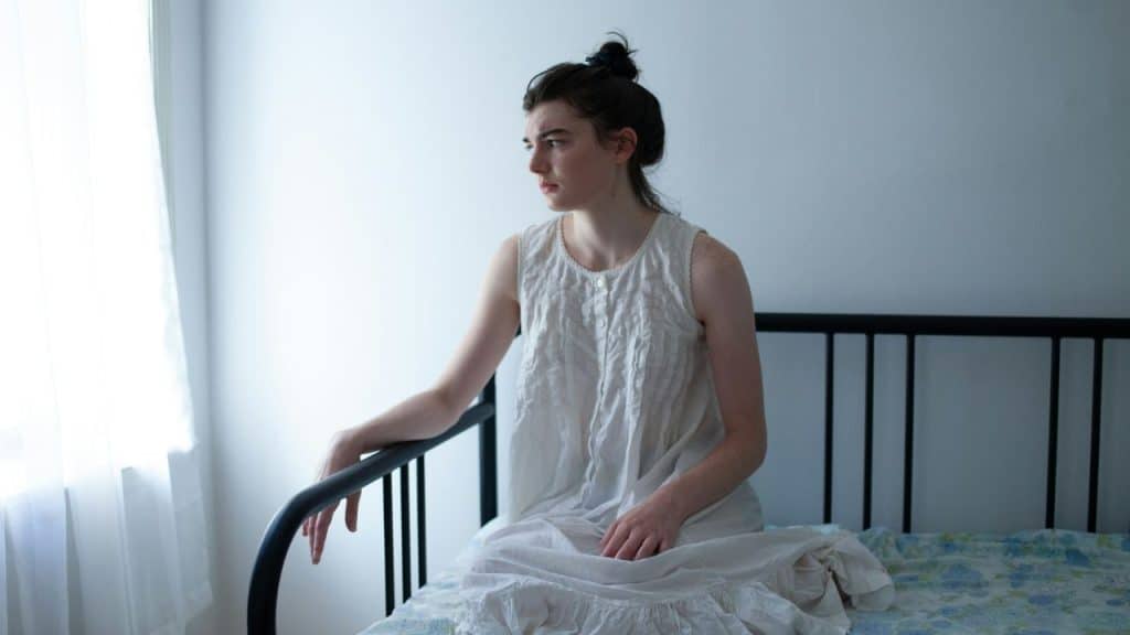 A woman in a white sleeveless nightgown sits on a bed and looks toward a window.