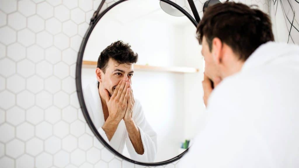 A man in a white robe looks at his reflection in a circular bathroom mirror.
