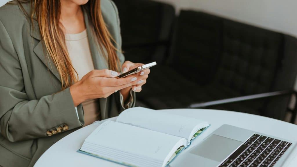 A person in a blazer uses a smartphone beside an open notebook and laptop.