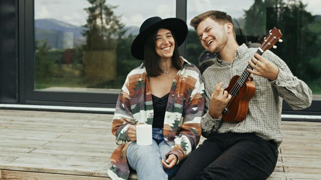 A laughing man plays a ukulele while sitting on a wooden deck next to a smiling woman.