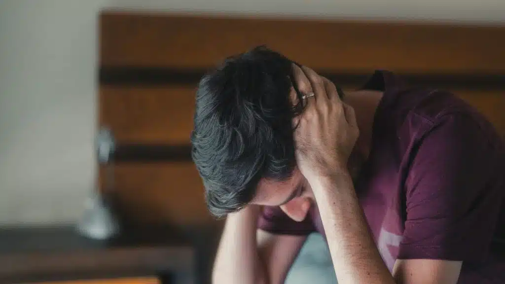 A close-up shot of a man in a purple shirt holding his head in his hands.