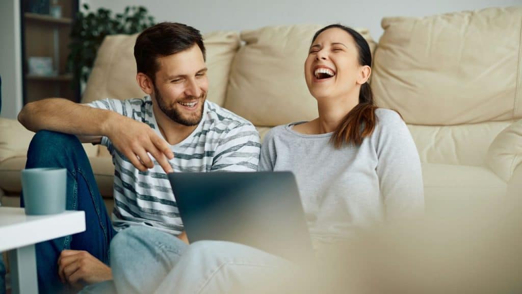 A laughing man and woman sit on the floor in front of a laptop computer.