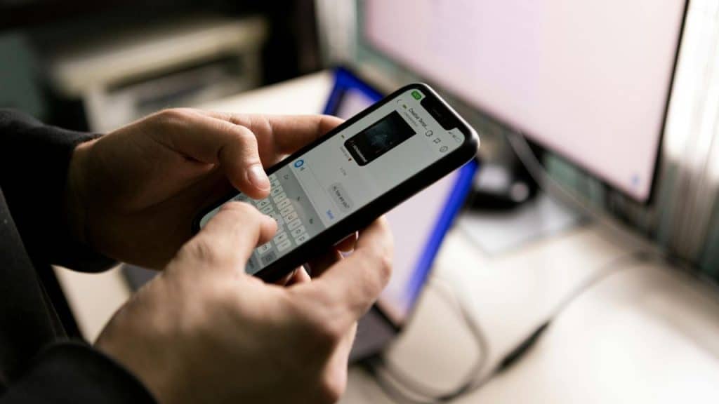 Close-up of hands typing a message on a smartphone in front of a computer monitor.