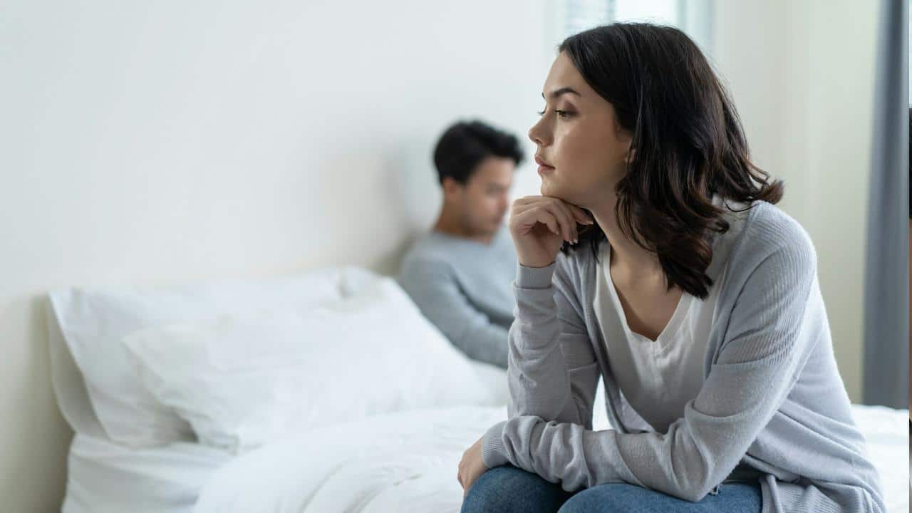 A woman looking sad while sitting on a bed as her partner sits on the bed in the background.