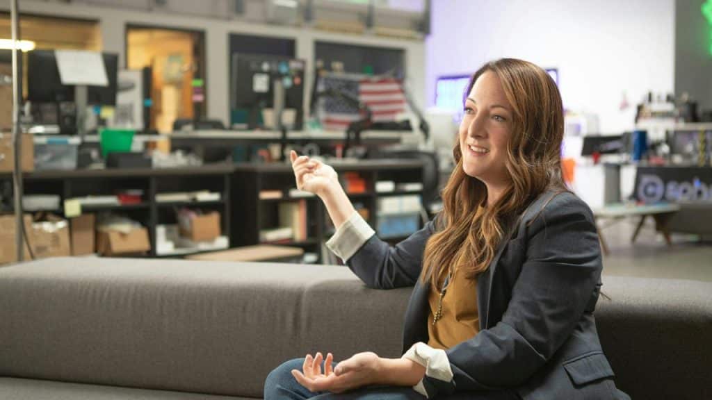 A woman sitting on a couch in an office, smiling while gesturing during a conversation.