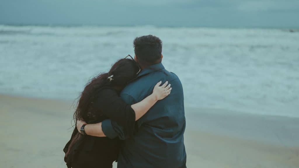A rearview shot of a couple standing on a sandy beach, looking out at the ocean waves under an overcast sky.
