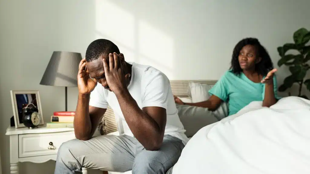 A man sits on the edge of a bed with his head in his hands, appearing stressed or upset, while a woman sits behind him on the bed, gesturing as if speaking.