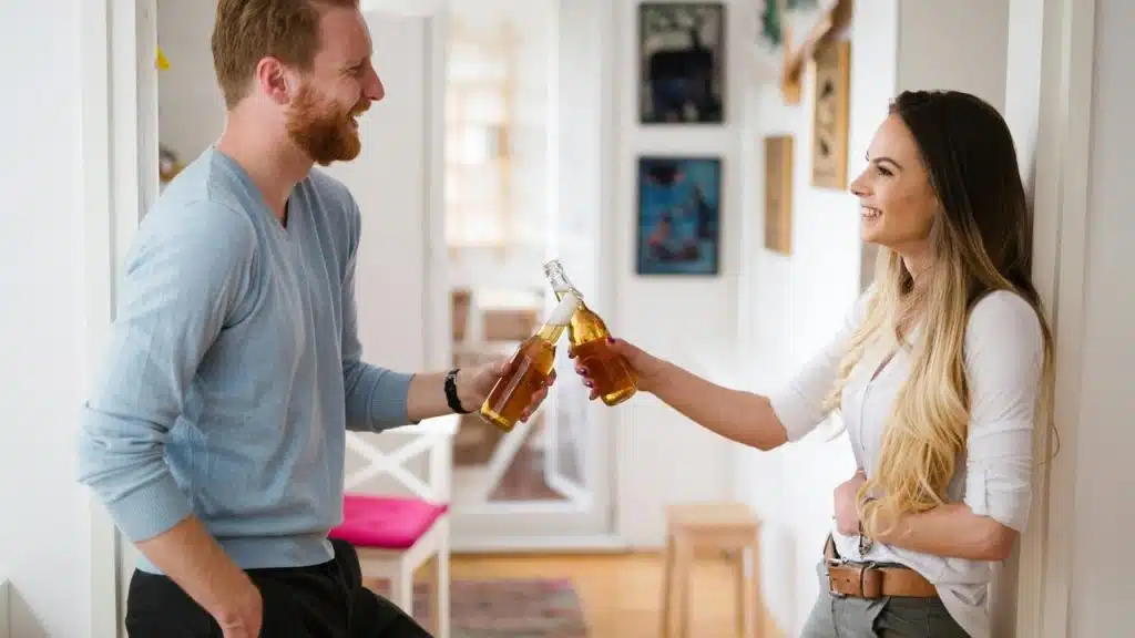 A man and woman smile while clinking beer bottles together indoors.
