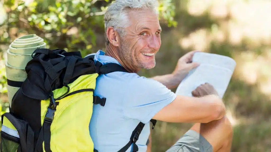 A smiling older man with a backpack and map sits outdoors in a sunlit area.