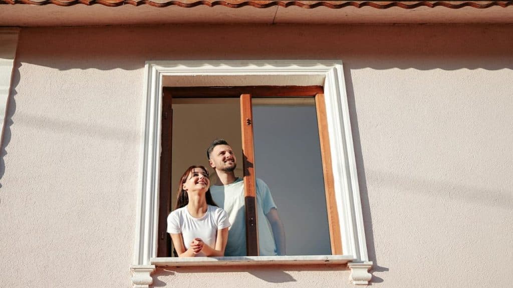 A man and woman look out of a window from inside a pink building.