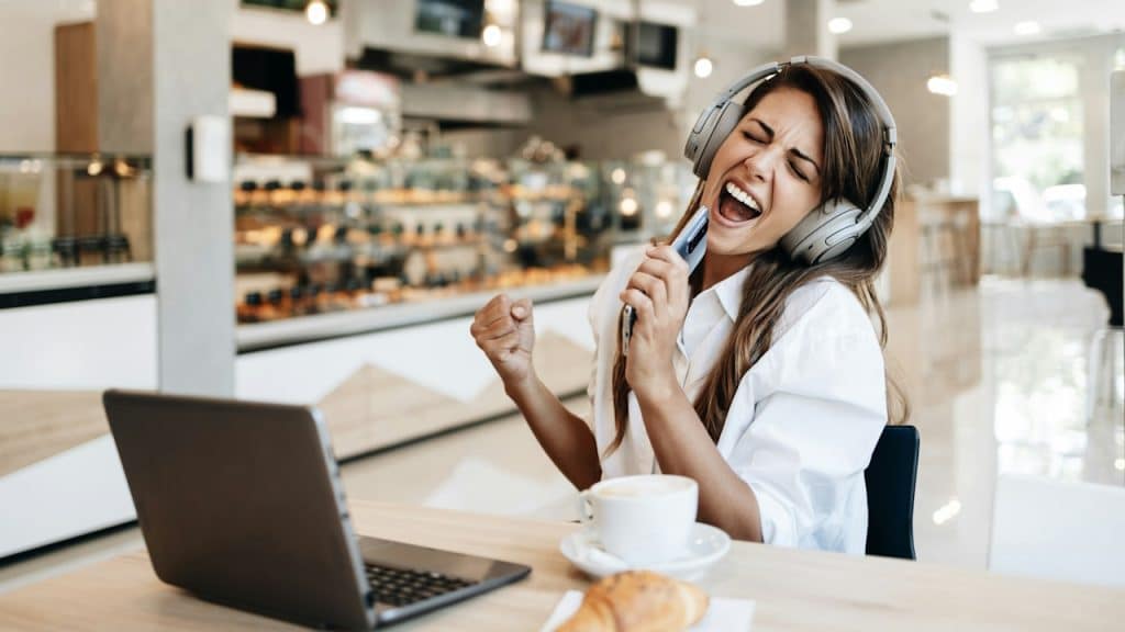 A woman is grooving while listening to music on her headphones while sitting on table on which her laptop is placed.