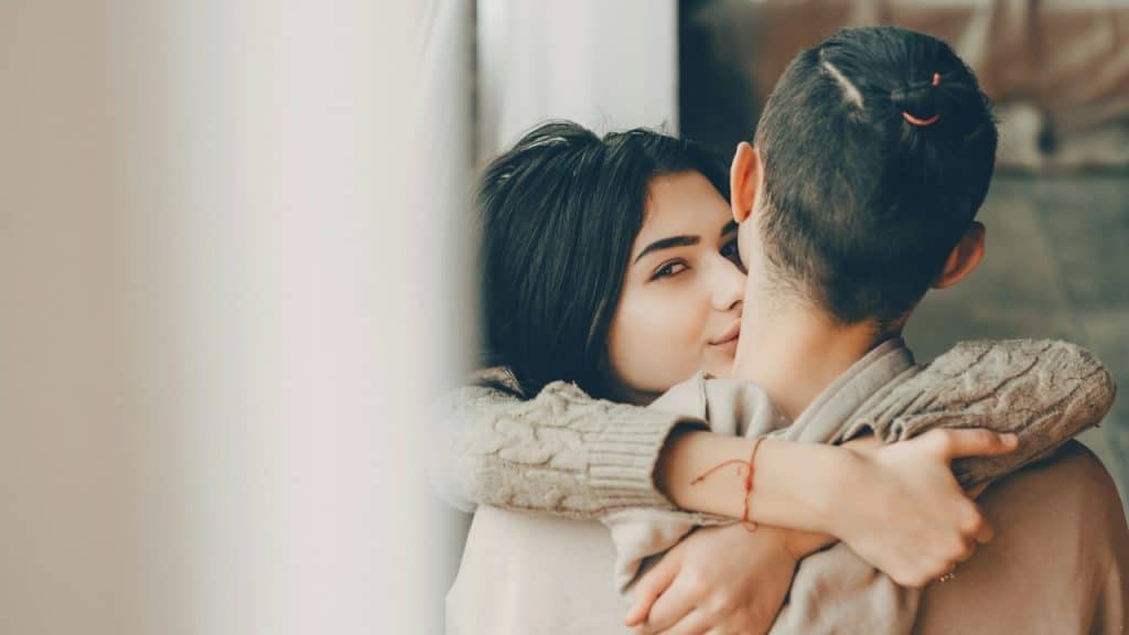 A woman is looking at the camera while hugging a man who has his hair tied in a ponytail.