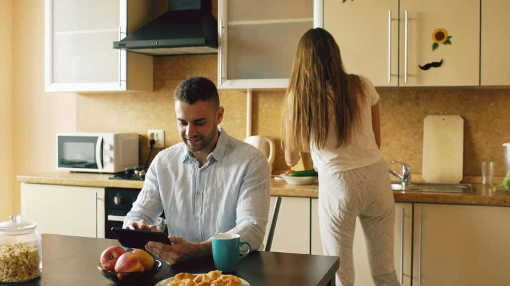 A man uses a tablet at a table while a woman stands with her back turned.