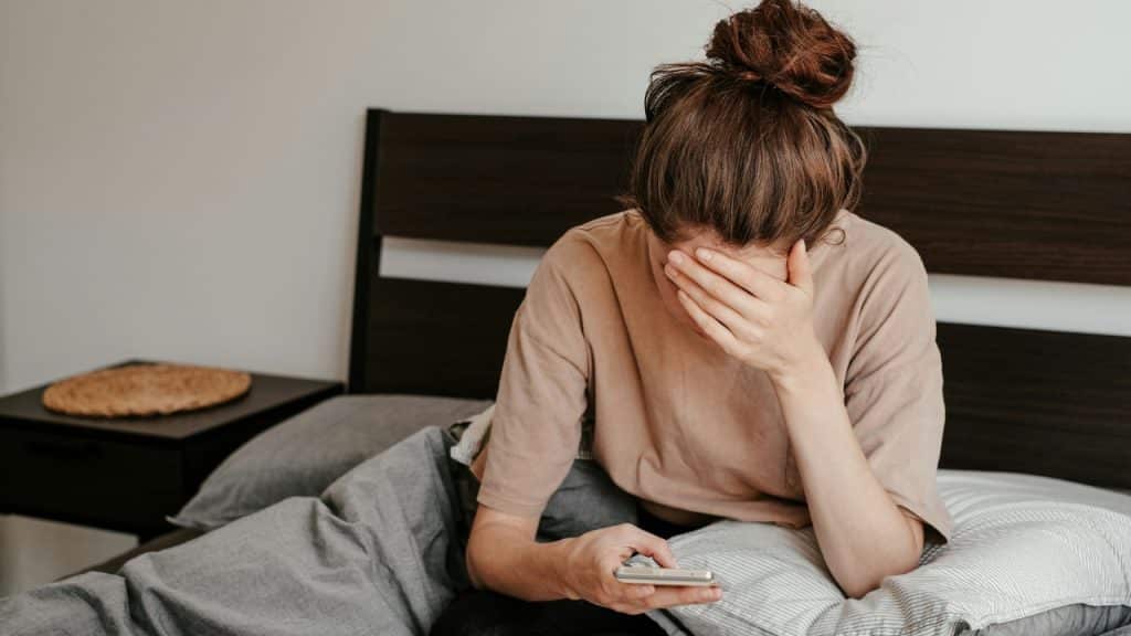 A woman sits in bed holding a phone while covering her face with her other hand.