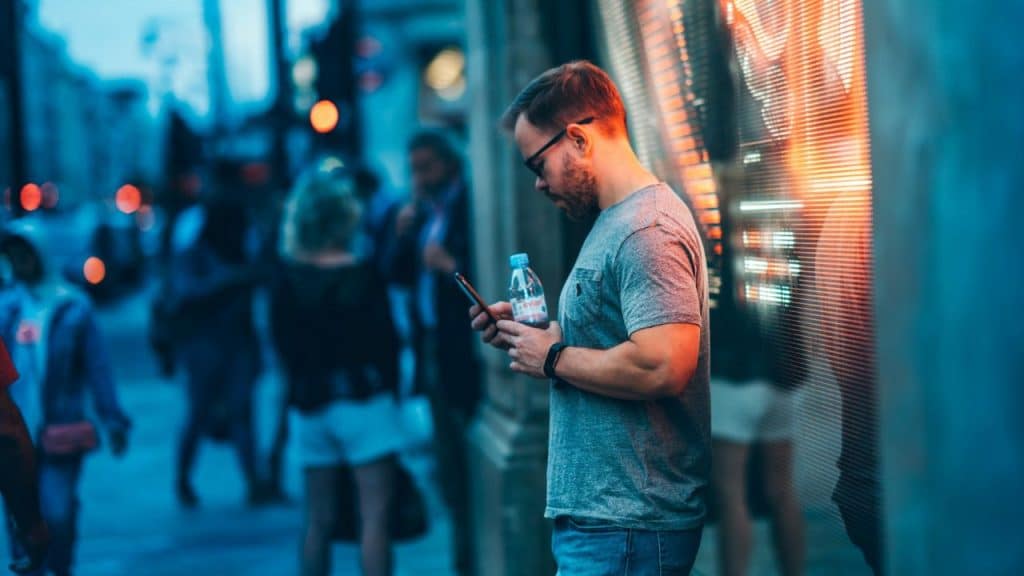 A man in a grey shirt looks at a phone on a crowded, blue-toned city street.