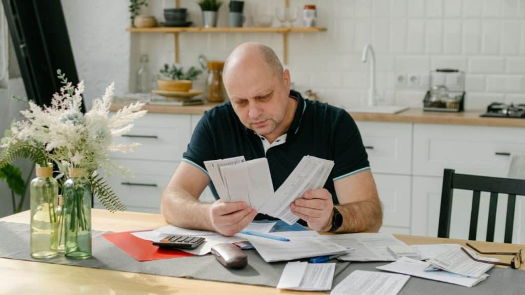 A bald man sitting at a kitchen table looking through several paper documents and bills.