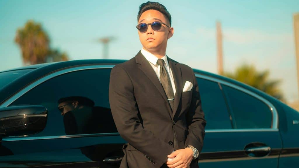 A man wearing sunglasses and a suit standing by a black car with hands clasped and looking to the side.