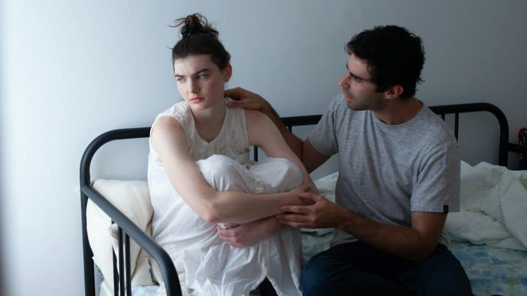 A man sits on a bed and places his hand on a woman’s shoulder.