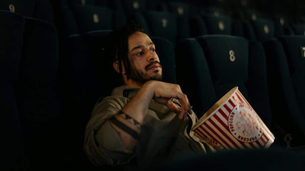 A man with dreadlocks sits alone in a dark theater, holding a striped popcorn bucket.