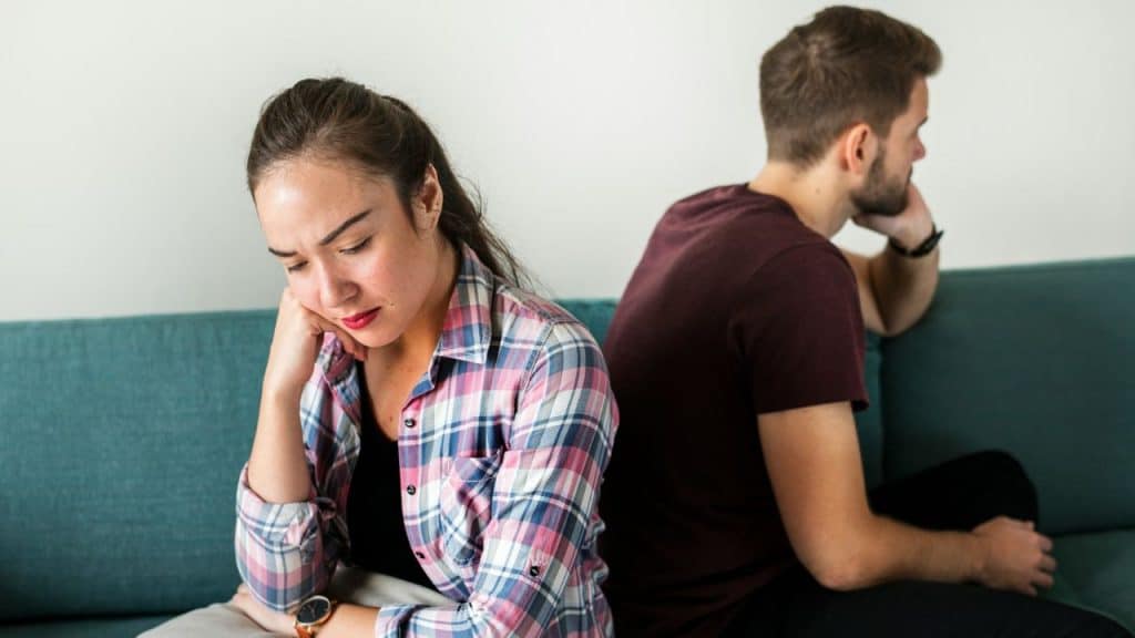 A man and woman sit back-to-back on a blue couch with downcast facial expressions.