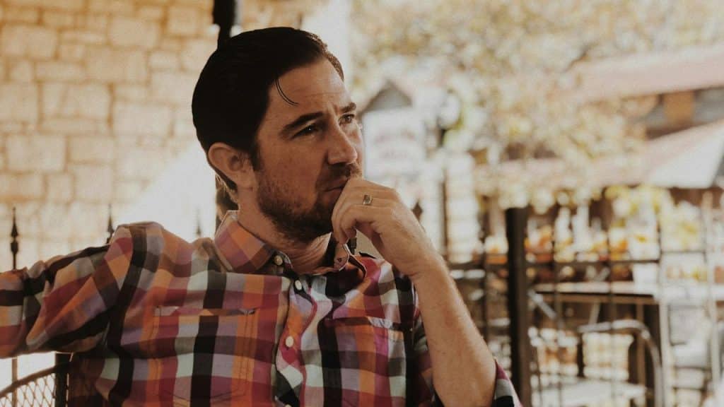 A man with dark hair and a beard wearing a plaid shirt sits outdoors thinking.