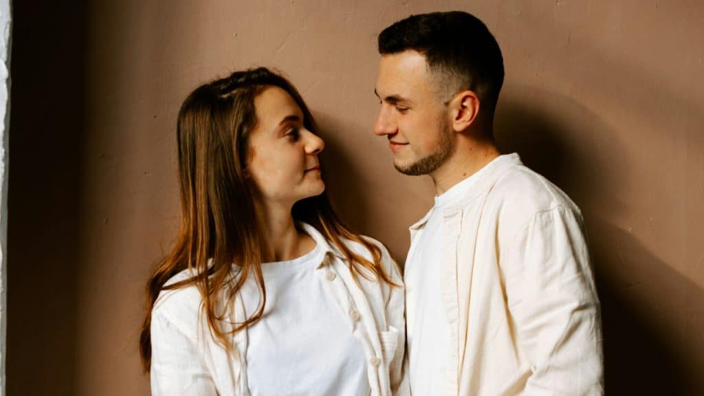 A man and woman in white shirts stand close together against a plain tan wall.